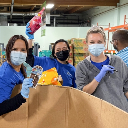 Three food bank volunteers wearing masks and placing donations in a bin.